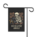 Garden flag with skeleton and flowers design and text 'DIG IN & STAY AWHILE' on a black background.