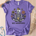 Heather purple t-shirt with a graphic of a cartoon skeleton sitting among flowers and bees that reads 'Bee Whisperer'.