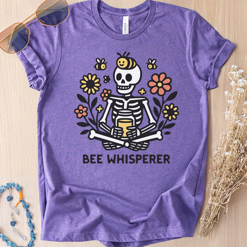 Heather purple t-shirt with a graphic of a cartoon skeleton sitting among flowers and bees that reads 'Bee Whisperer'.