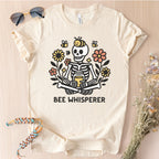 Bella Canvas 3001 in Natural 'Bee Whisperer' t-shirt featuring a graphic of a cartoon skeleton sitting with bees and flowers.
