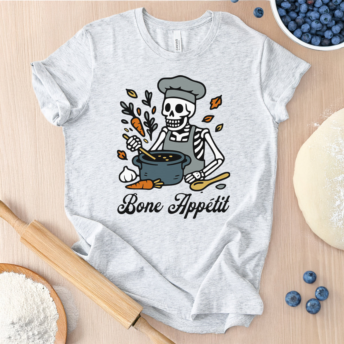 White t-shirt with a skeleton chef graphic and 'Bone Appetit' text on a wooden surface with baking items.