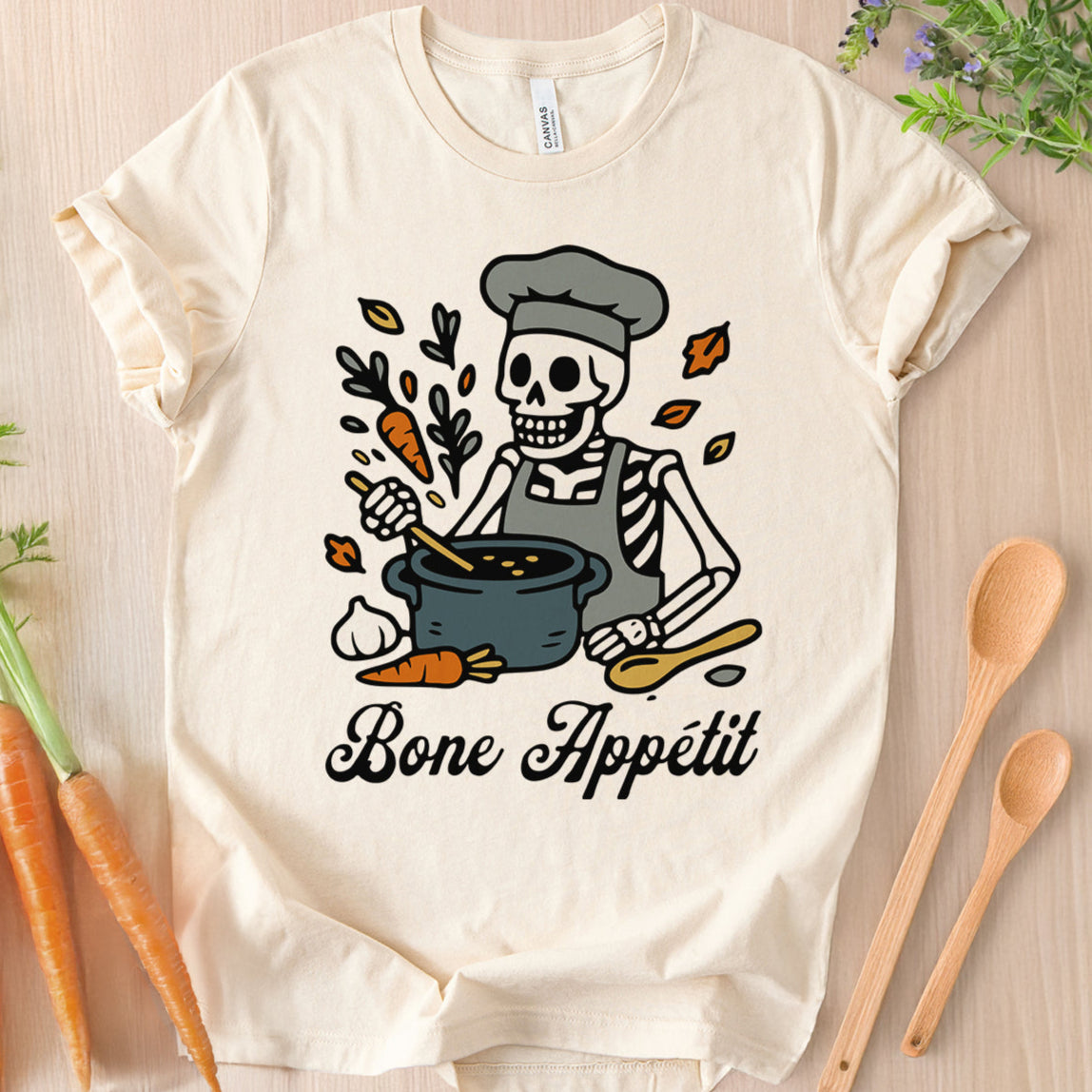 Flat lay of a Natural colored Bella Canvas 3001 t-shirt with a cartoon skeleton chef stirring a pot, with the text 'Bone Appétit'.