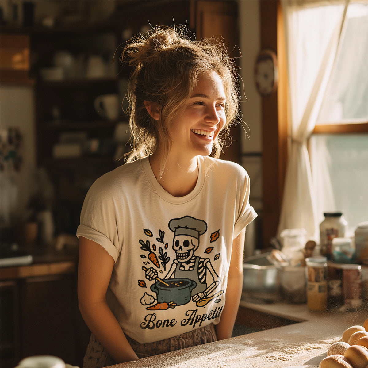 Woman smiling while wearing the Natural colored 'Bone Appétit' skeleton chef t-shirt in a warm, sunlit kitchen.