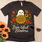 Black t-shirt with a ghost and floral design labeled 'Boo-tiful Blooms' on a wooden surface with sunflowers and sunglasses.