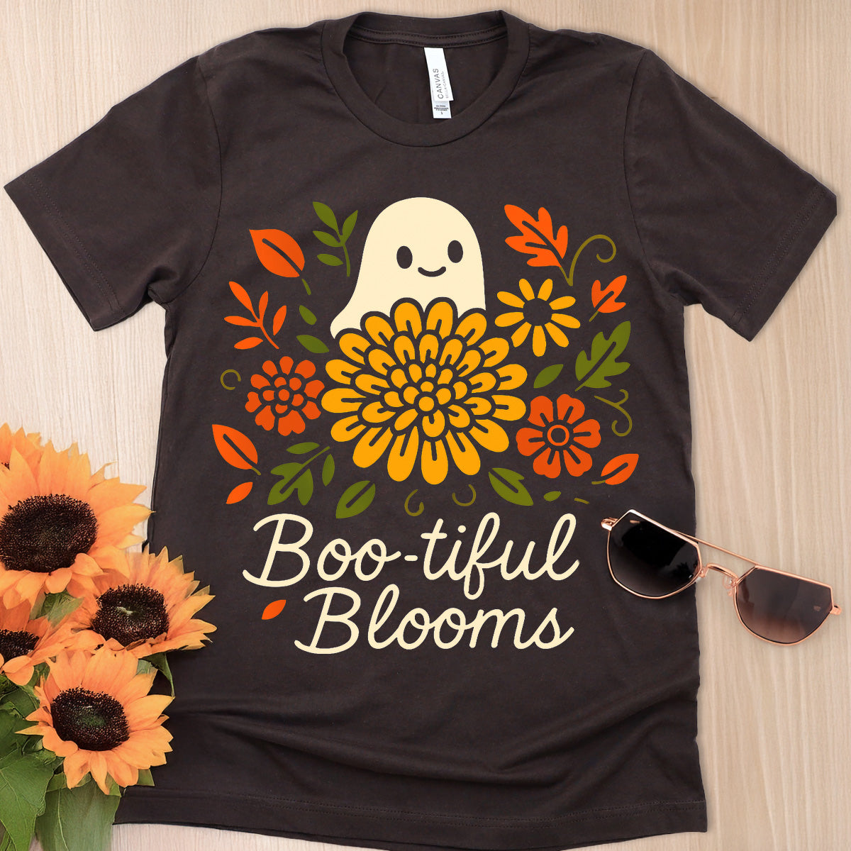Black t-shirt with a ghost and floral design labeled 'Boo-tiful Blooms' on a wooden surface with sunflowers and sunglasses.