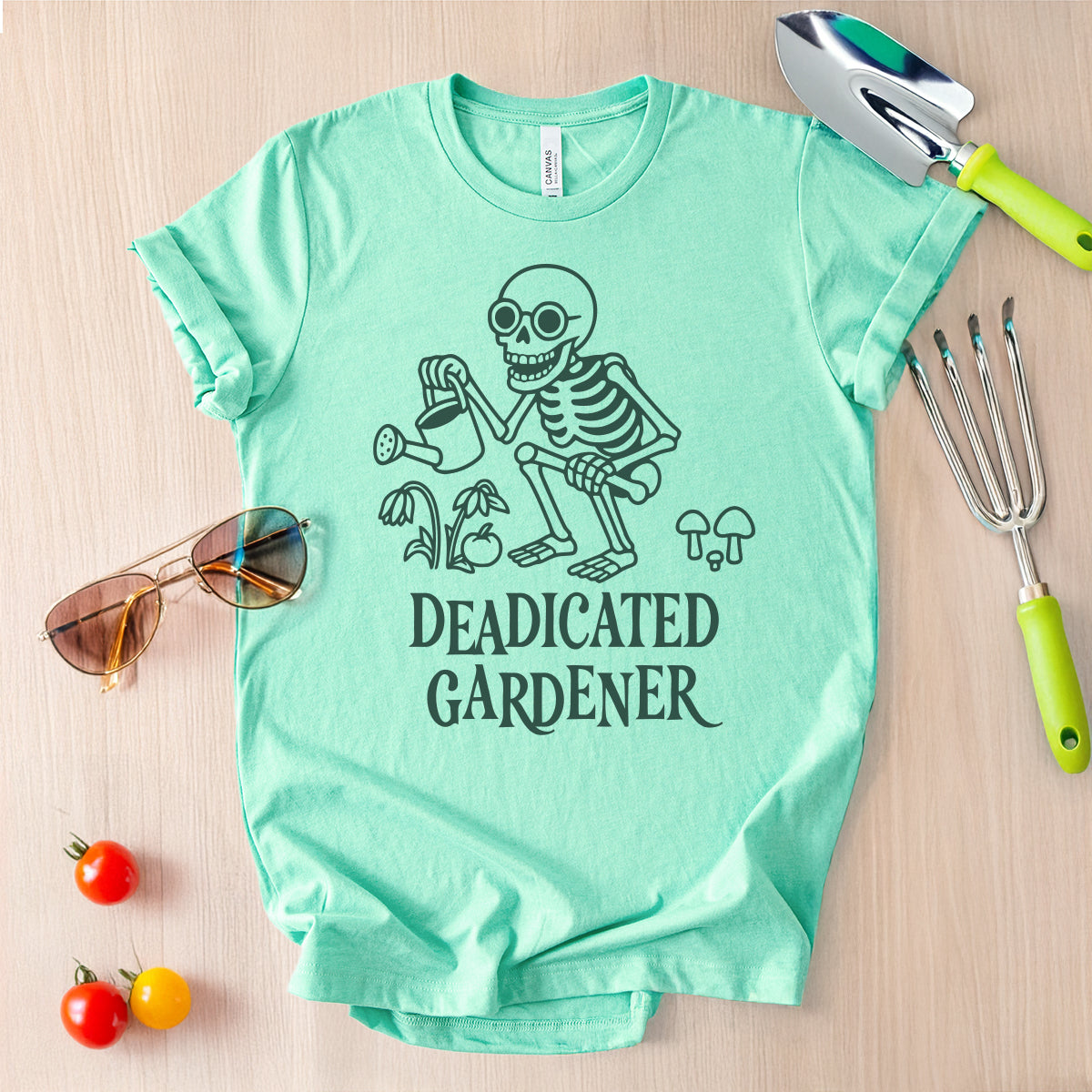 Mint green t-shirt with skeleton graphic and 'DEADICATED GARDENER' text, sunglasses, and gardening tools on a wooden surface.