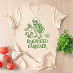 Beige t-shirt with green skeleton graphic and 'DEADICATED GARDENER' text on a wooden surface with tomatoes and plants.