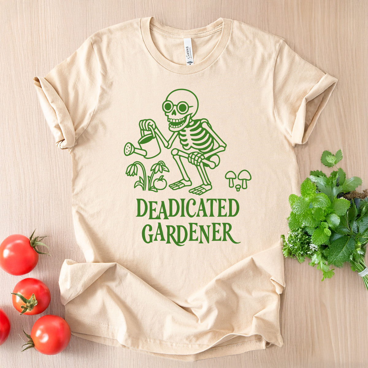 Beige t-shirt with green skeleton graphic and 'DEADICATED GARDENER' text on a wooden surface with tomatoes and plants.