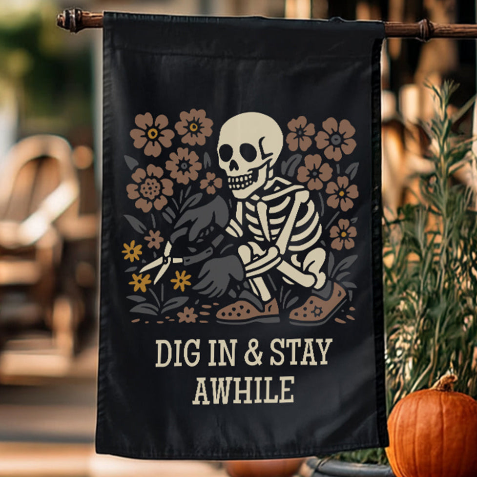 Decorative flag with skeleton and flowers design, featuring text 'DIG IN & STAY AWHILE', hanging outdoors.