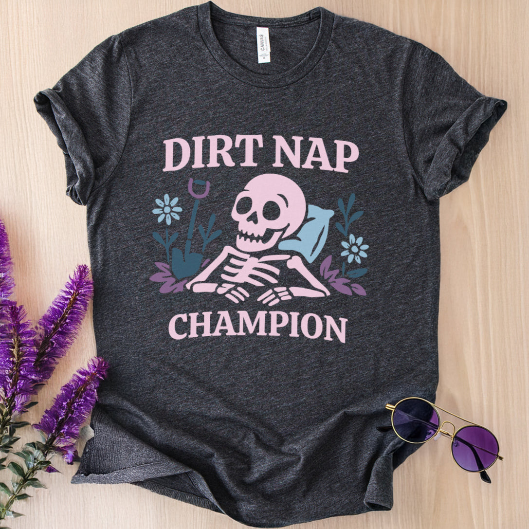 Dark gray t-shirt with skeleton design and text 'Dirt Nap Champion' on a wooden surface with purple flowers and sunglasses.
