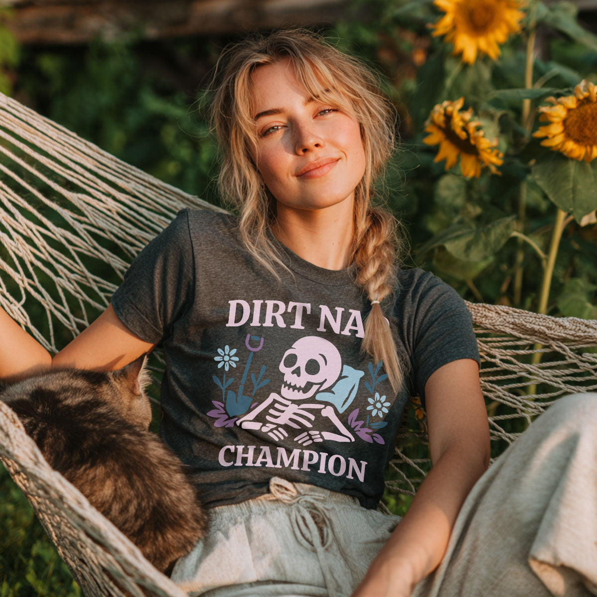Woman wearing a 'Dirt N A Champion' t-shirt sitting in a hammock with sunflowers in the background