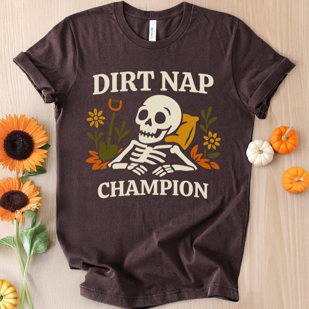 Brown t-shirt with 'Dirt Nap Champion' graphic on a wooden surface with decorative items.