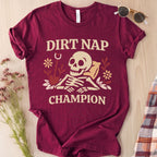 Maroon t-shirt with 'Dirt Nap Champion' graphic on a wooden surface with sunglasses and flowers.