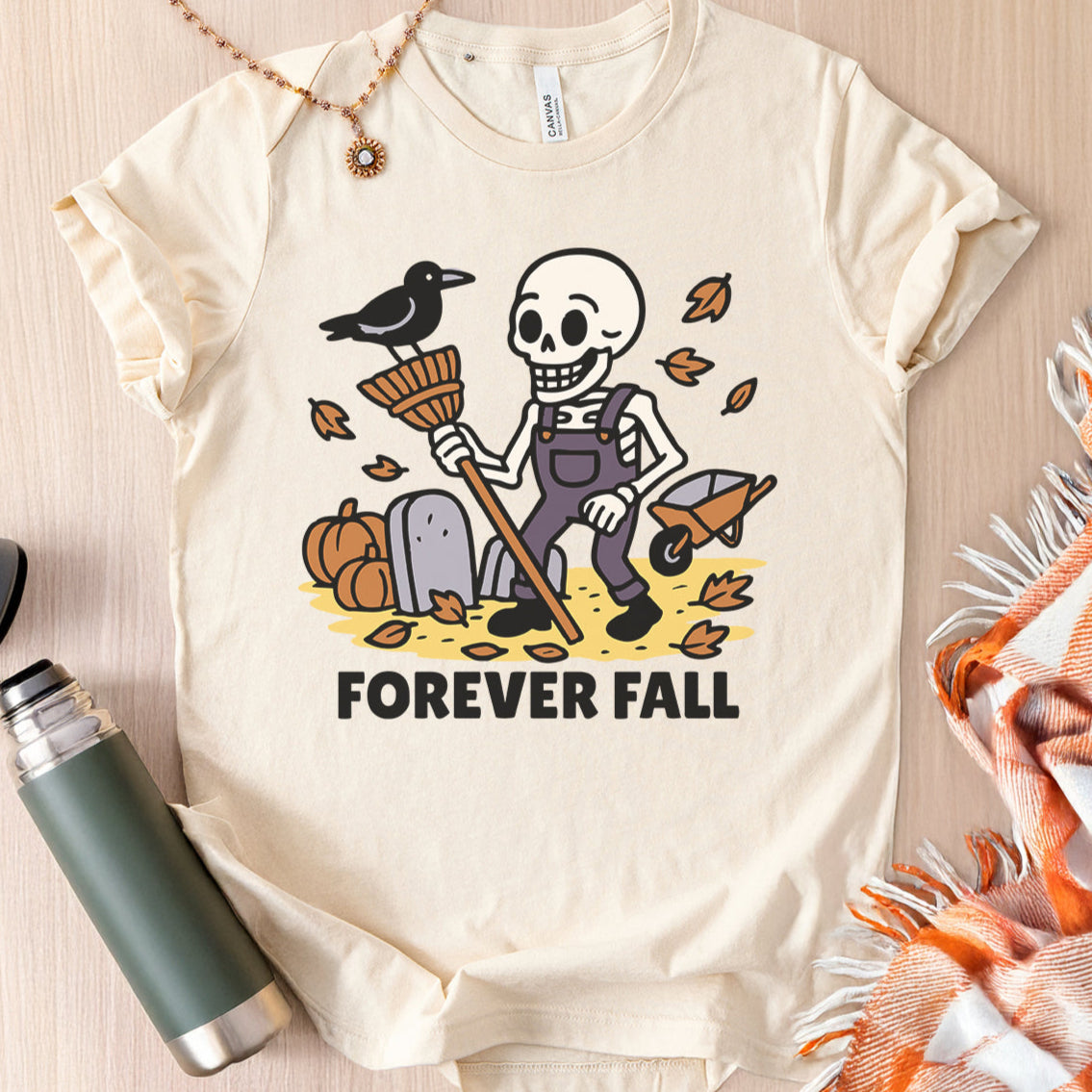Autumn-themed flat lay of a natural colored Bella Canvas 3001 tshirt with a cartoon skeleton raking leaves that reads 'Forever Fall'.