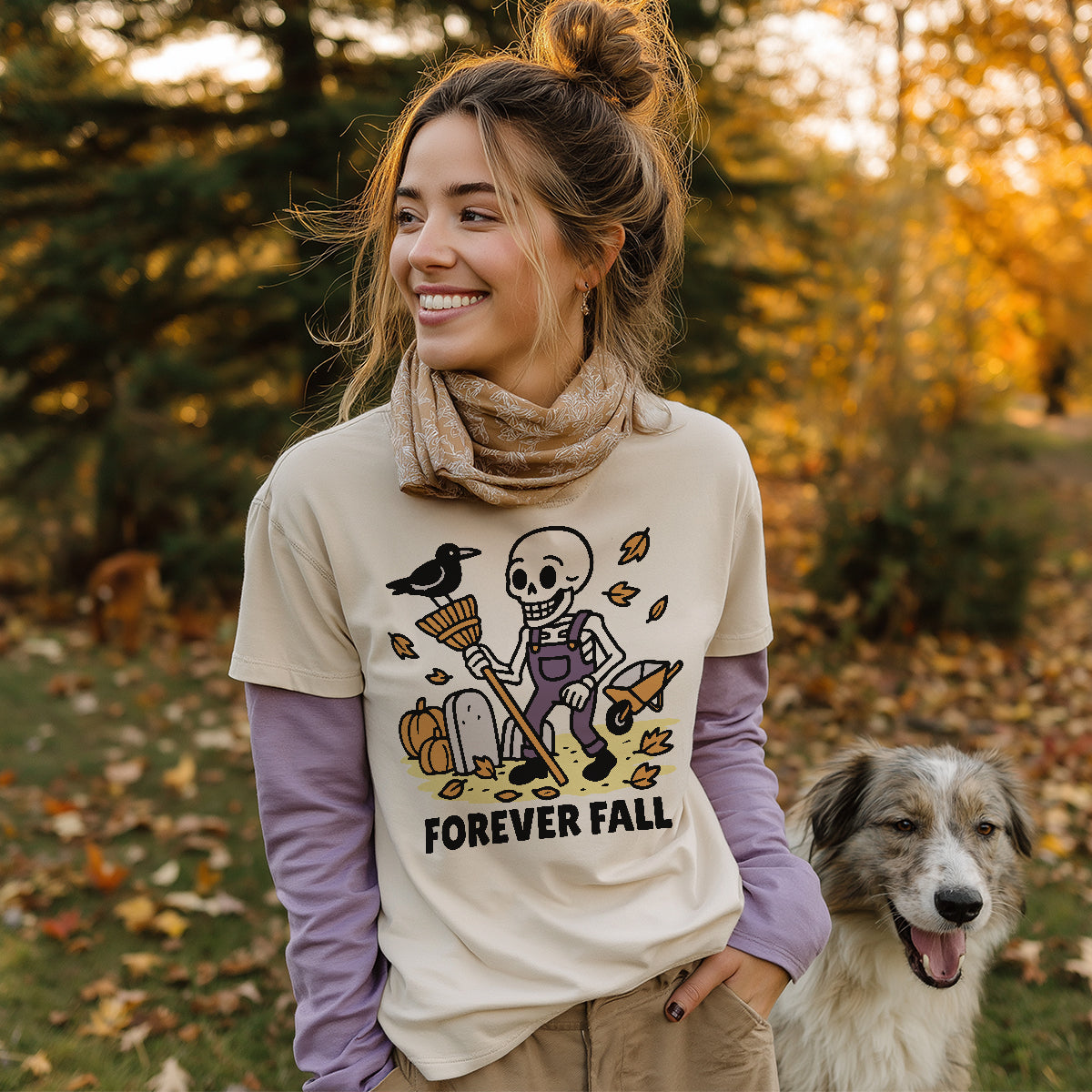 Model wearing the cream 'Forever Fall' skeleton t-shirt layered over a long-sleeve shirt, smiling outside in an autumn park with her dog.