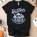 Halloween-themed flat lay of a black t-shirt featuring a graphic of purple mushroom houses that reads 'Fueled by Coffee & Mischief'.
