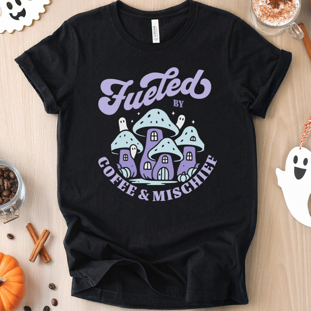 Halloween-themed flat lay of a black t-shirt featuring a graphic of purple mushroom houses that reads 'Fueled by Coffee & Mischief'.