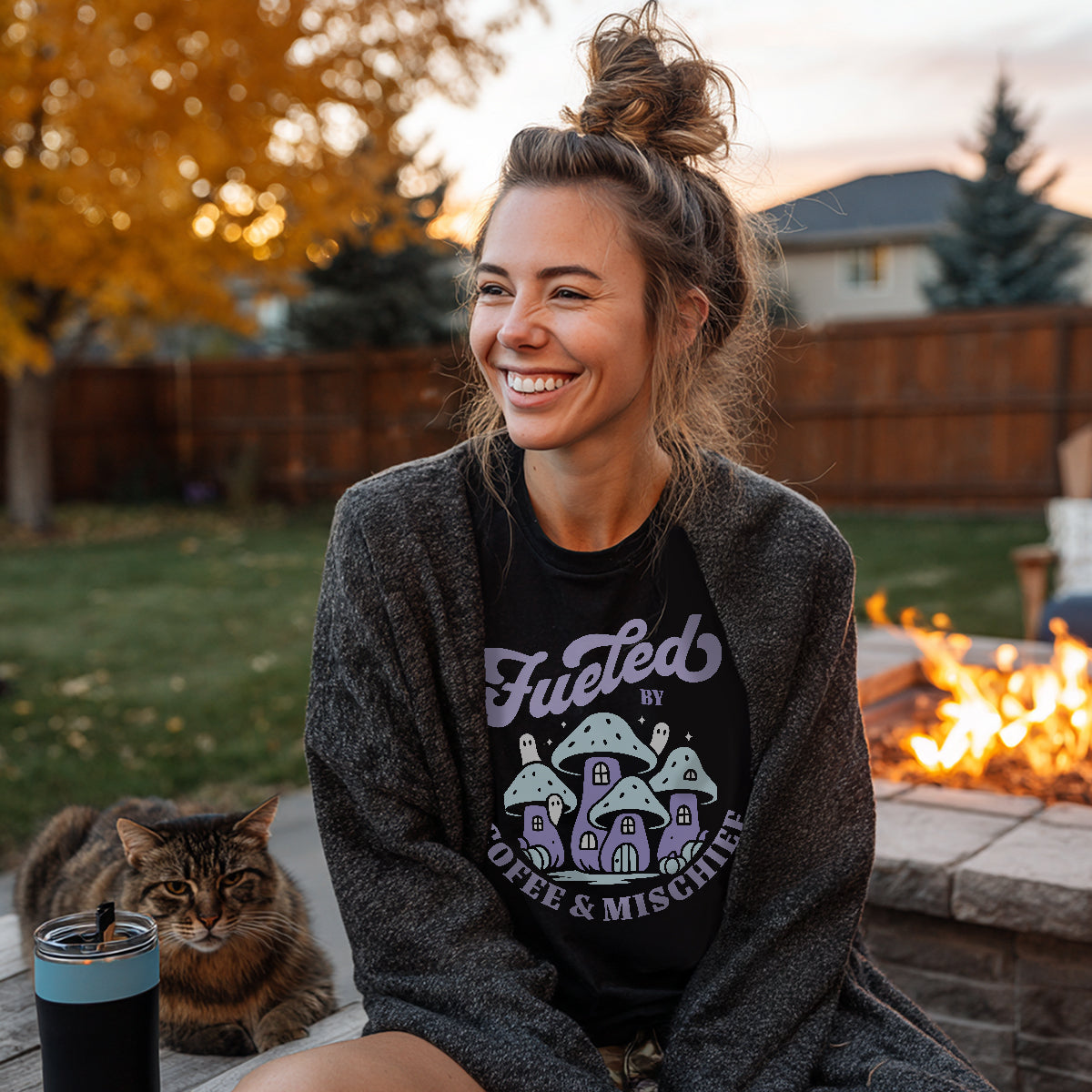 Model smiling while wearing the black 'Fueled by Coffee & Mischief' mushroom shirt, sitting outside by a fire pit with a cat.
