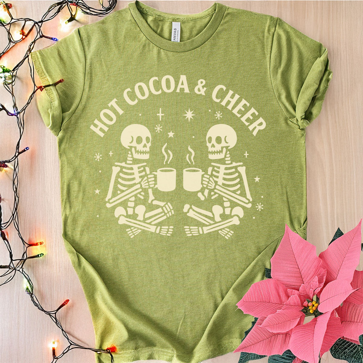 Green t-shirt with skeleton design and 'Hot Cocoa & Cheer' text on a wooden surface with Christmas lights and a pink flower.