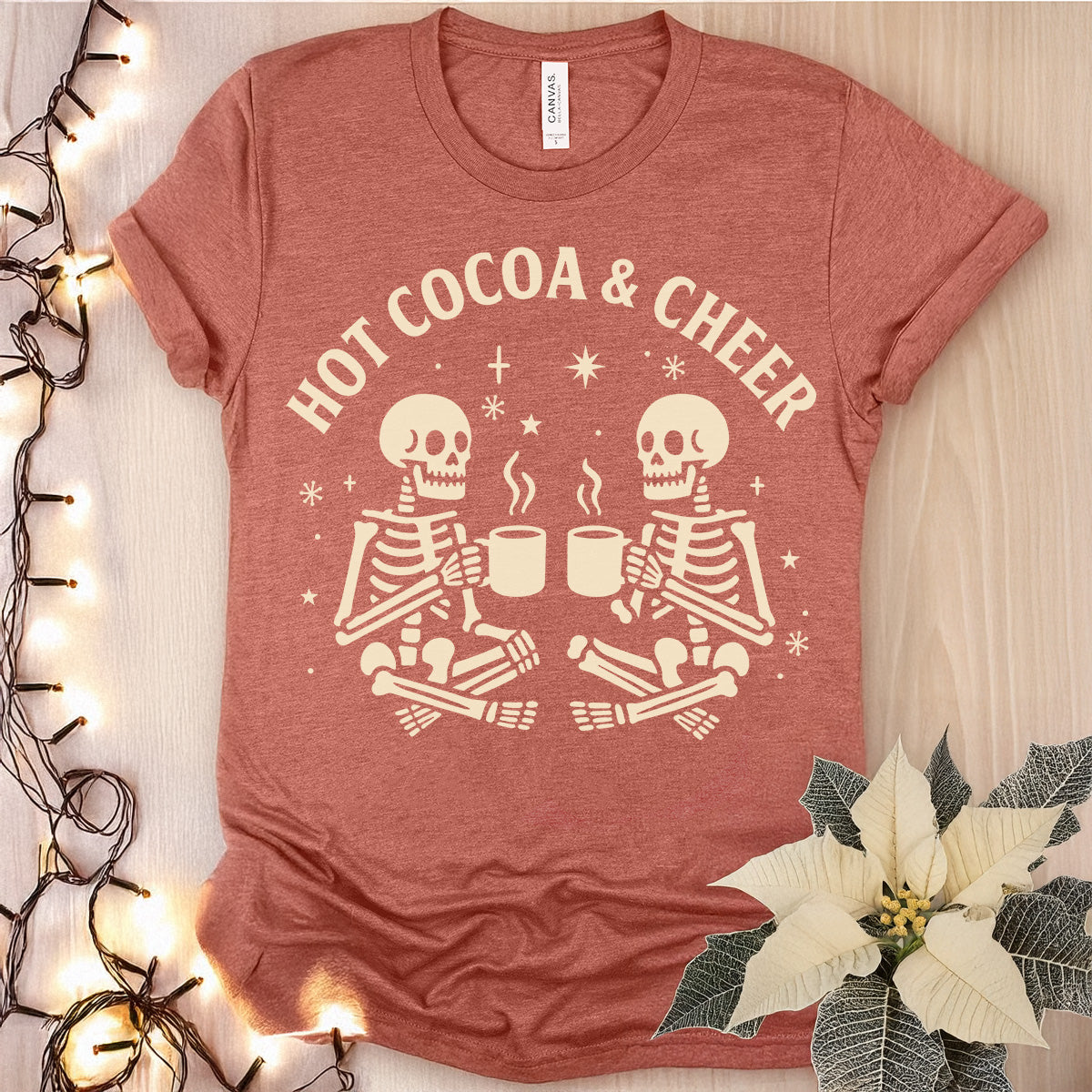 Red t-shirt with 'Hot Cocoa & Cheer' and skeleton design on a wooden surface with lights and a poinsettia.