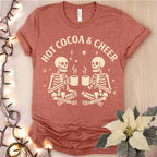Red t-shirt with 'Hot Cocoa & Cheer' and skeleton design on a wooden surface with lights and a poinsettia.