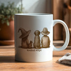 White mug with a design of three figures and the text 'kindred magic' on a wooden surface.
