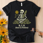 Flat lay of a black t-shirt featuring a cartoon skeleton on a pile of pickles with the text 'R.I.P. (Rest in Pickles)'.