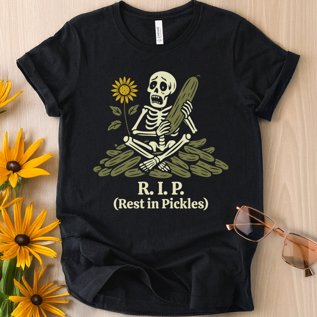 Flat lay of a black t-shirt featuring a cartoon skeleton on a pile of pickles with the text 'R.I.P. (Rest in Pickles)'.