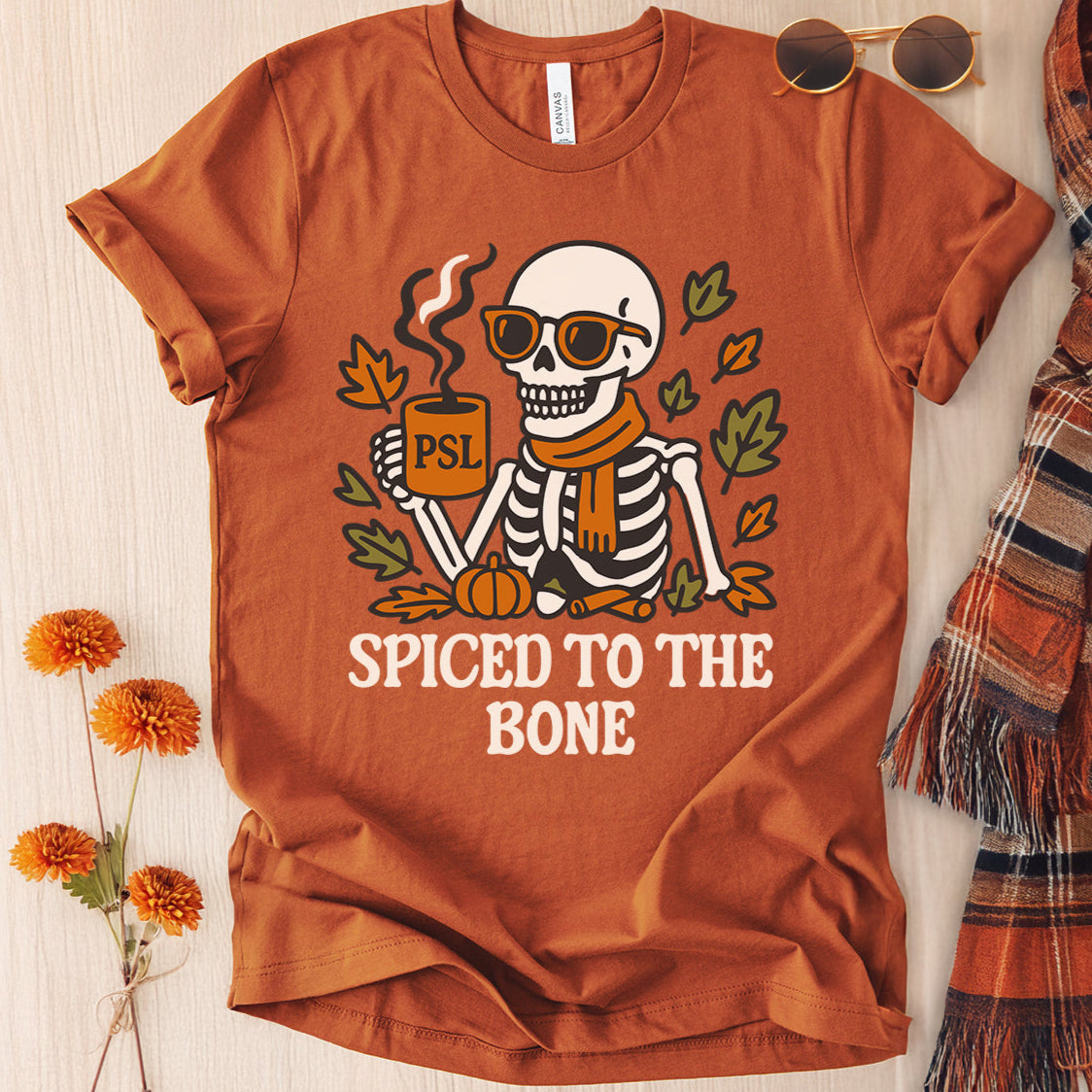 Brown t-shirt with skeleton design and 'Spiced to the Bone' text, surrounded by autumn-themed items.