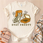 Beige t-shirt with skeleton graphic and 'Stay Frosty' text, surrounded by winter accessories on a light wooden surface.