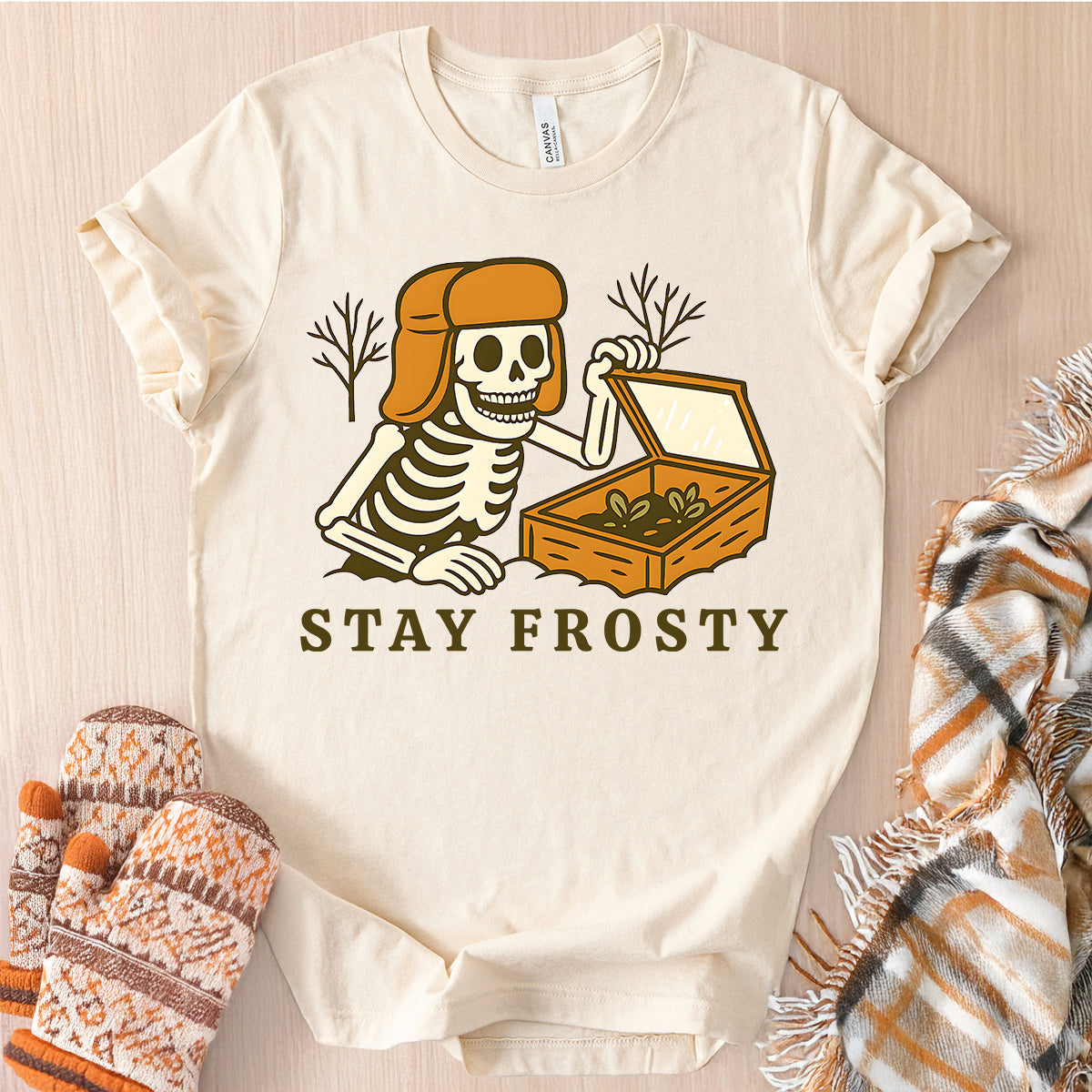 Beige t-shirt with skeleton graphic and 'Stay Frosty' text, surrounded by winter accessories on a light wooden surface.