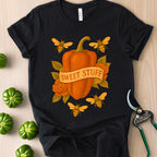 Black t-shirt with a pumpkin and bees design on a wooden surface with green peppers and shears.