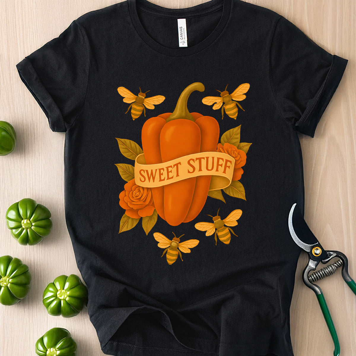 Black t-shirt with a pumpkin and bees design on a wooden surface with green peppers and shears.