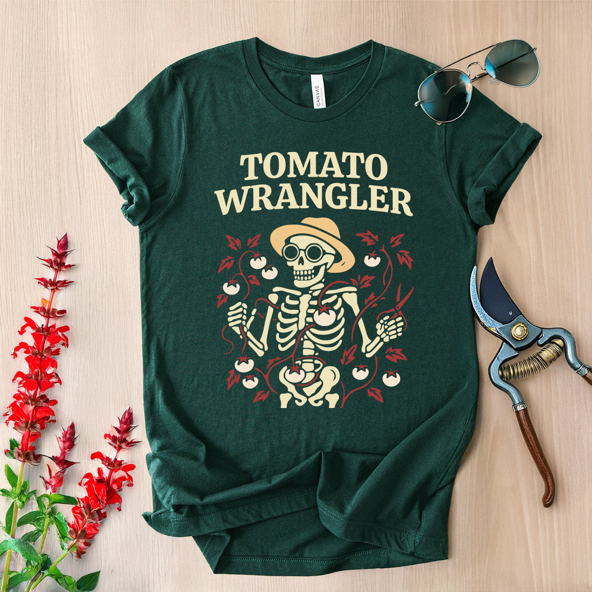 Green t-shirt with 'Tomato Wrangler' design on a wooden surface with gardening tools and flowers.