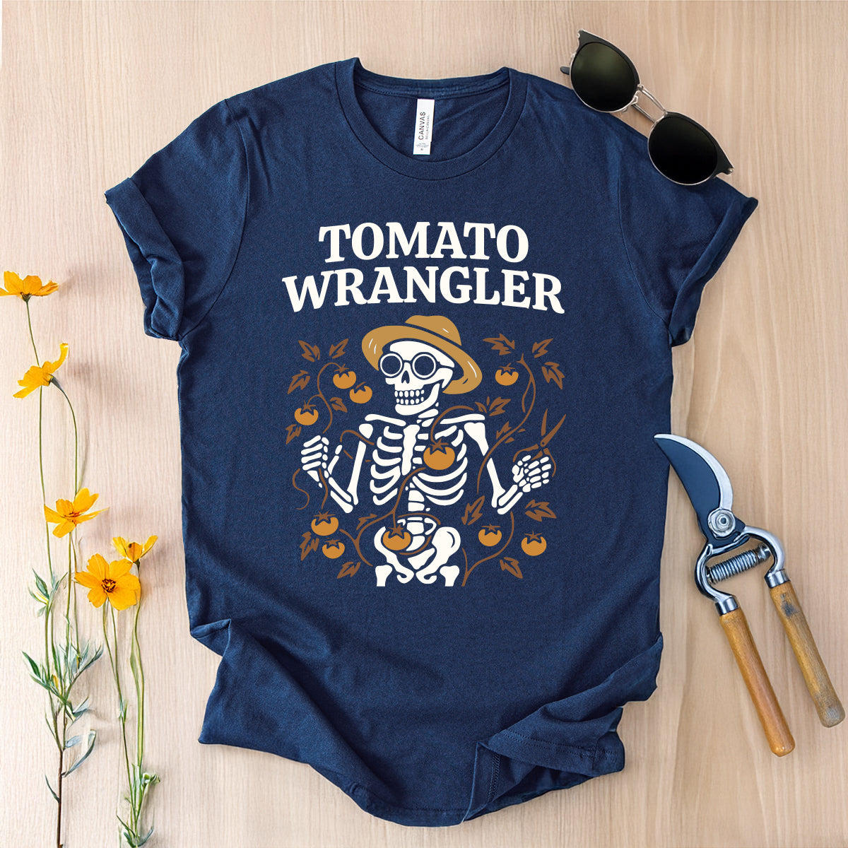Navy blue t-shirt with skeleton graphic and 'Tomato Wrangler' text on a wooden surface with sunglasses and garden tools.