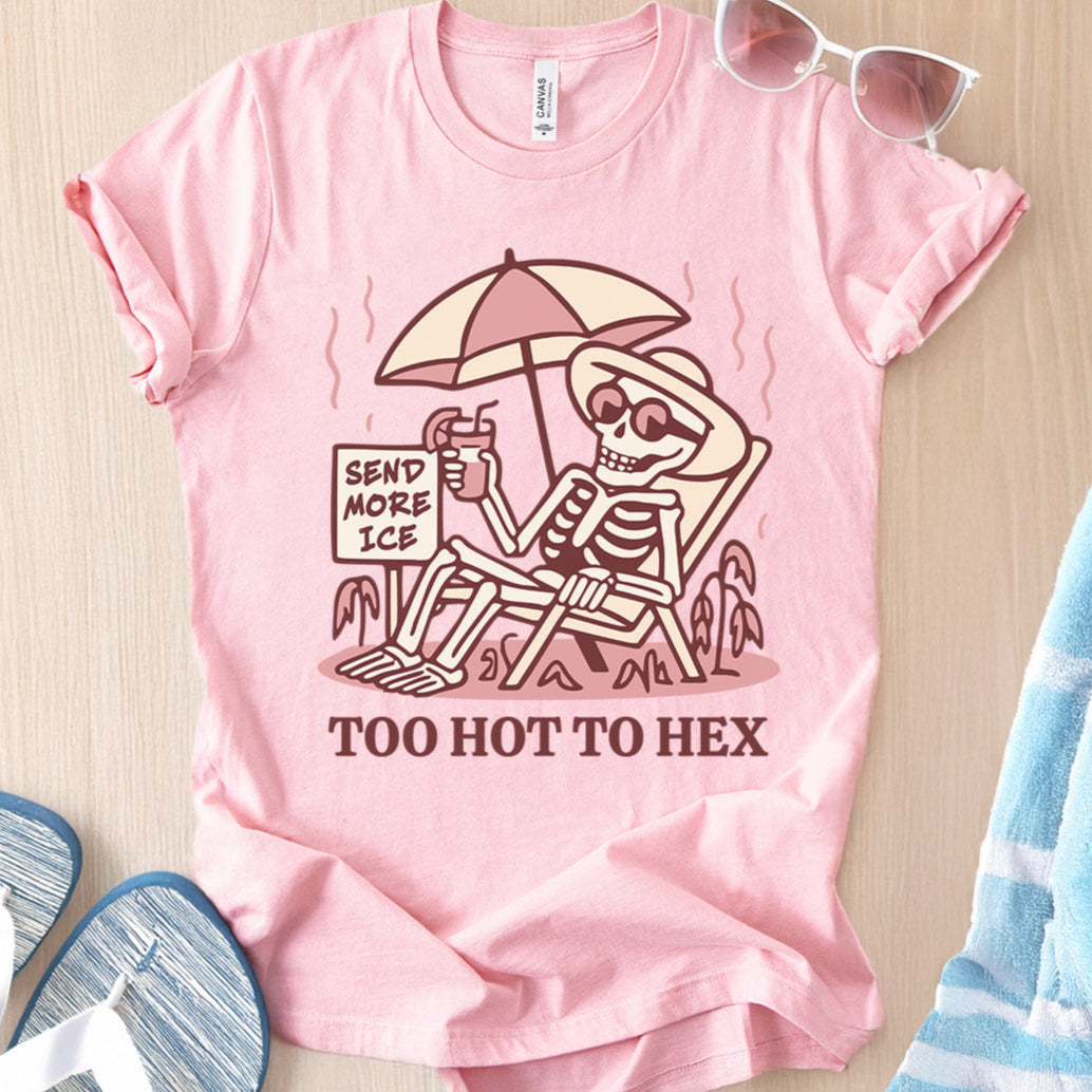 Pink t-shirt with skeleton design and text 'Send More Ice, Too Hot To Hex' on a wooden surface.