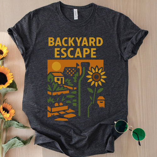 Dark gray t-shirt with 'Backyard Escape' graphic design on a wooden surface with sunflowers and sunglasses.