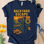 Navy blue t-shirt with 'Backyard Escape' graphic design on a wooden surface with gardening tools and flowers.