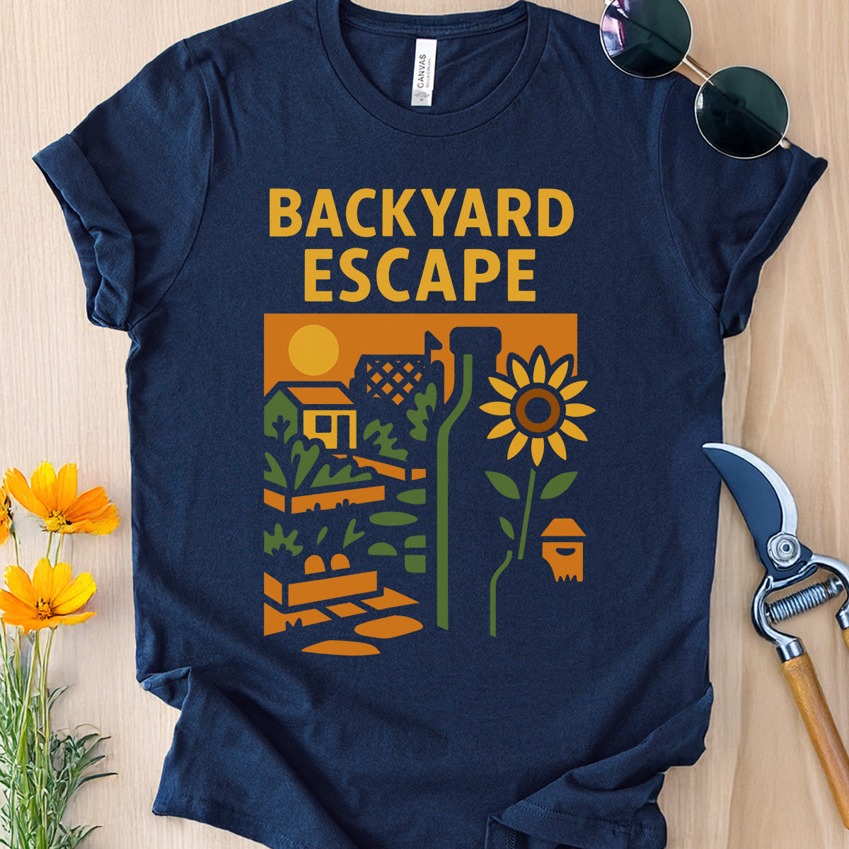 Navy blue t-shirt with 'Backyard Escape' graphic design on a wooden surface with gardening tools and flowers.