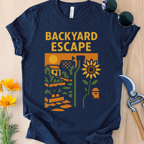 Navy blue t-shirt with 'Backyard Escape' graphic design on a wooden surface with gardening tools and flowers.