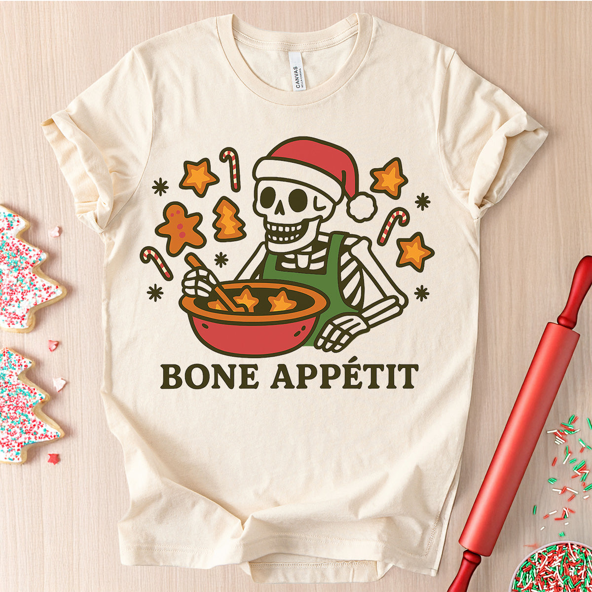 Beige t-shirt with a skeleton cooking design and 'Bone Appétit' text, surrounded by Christmas cookies and a rolling pin.