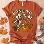 Orange t-shirt with skeleton design and 'Bone to Rake' text on a wooden surface with fall leaves.