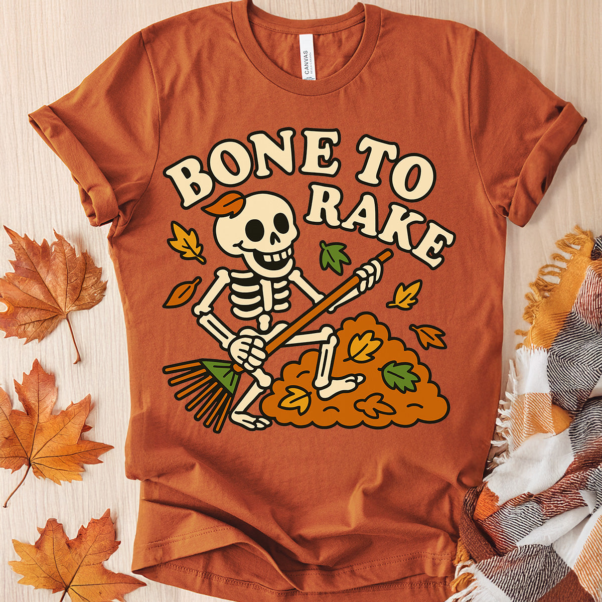 Orange t-shirt with skeleton design and 'Bone to Rake' text on a wooden surface with fall leaves.