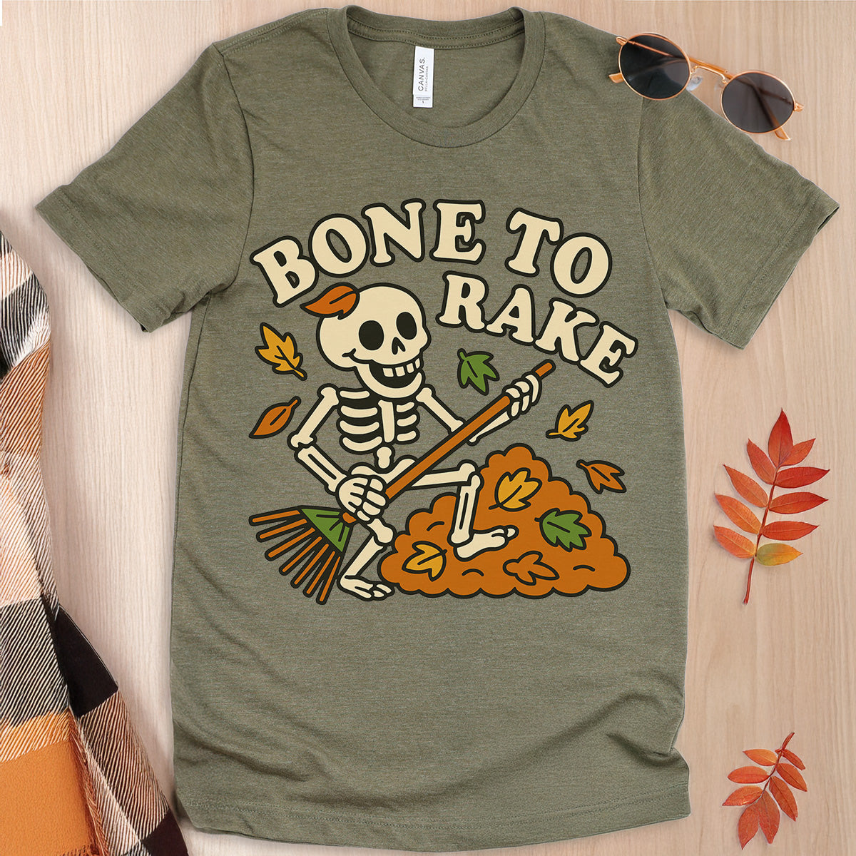 Green t-shirt with skeleton design and 'Bone to Rake' text on a wooden surface.