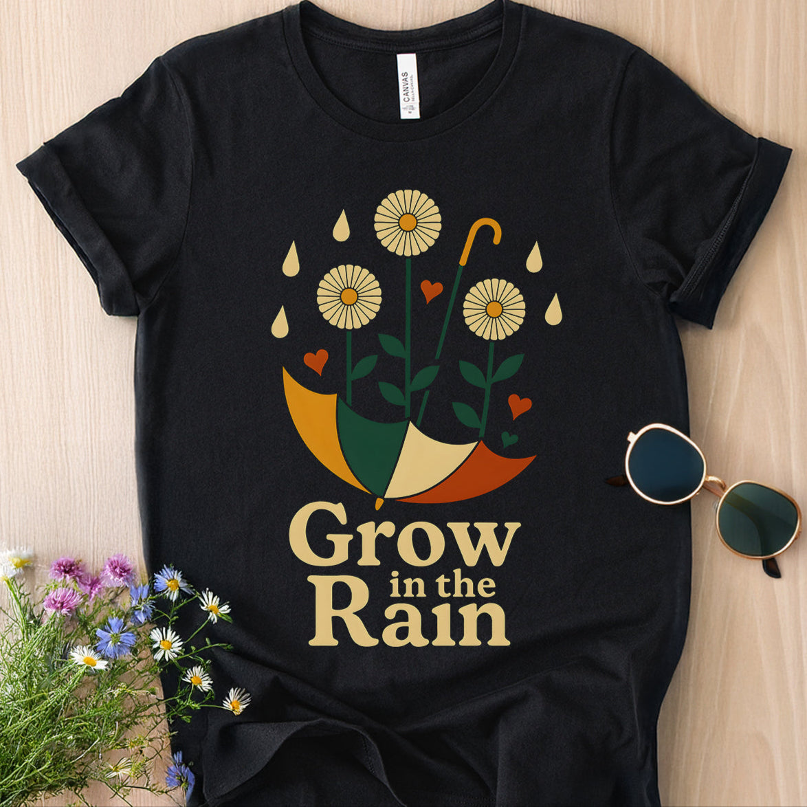 Black t-shirt with 'Grow in the Rain' design on a wooden surface with sunglasses and flowers.
