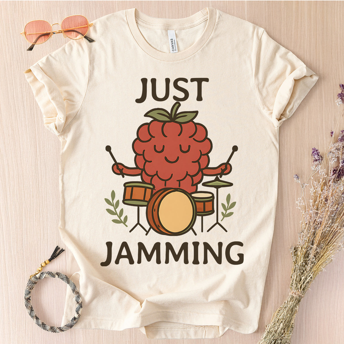 Beige t-shirt with a raspberry playing drums graphic and 'Just Jamming' text on a light wooden surface.