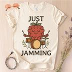 Beige t-shirt with a raspberry playing drums graphic and 'Just Jamming' text on a light wooden surface.