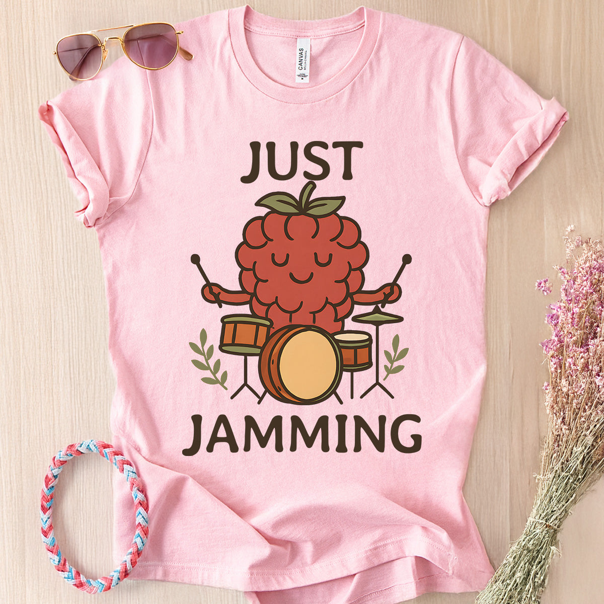 Pink t-shirt with a raspberry playing drums graphic and 'Just Jamming' text, sunglasses, and headband on a wooden surface.