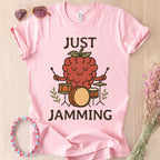 Pink t-shirt with a raspberry playing drums graphic and 'Just Jamming' text, sunglasses, and headband on a wooden surface.