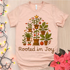 Pink t-shirt with plant design and 'Rooted in Joy' text on a wooden surface with decorative lights and flowers.